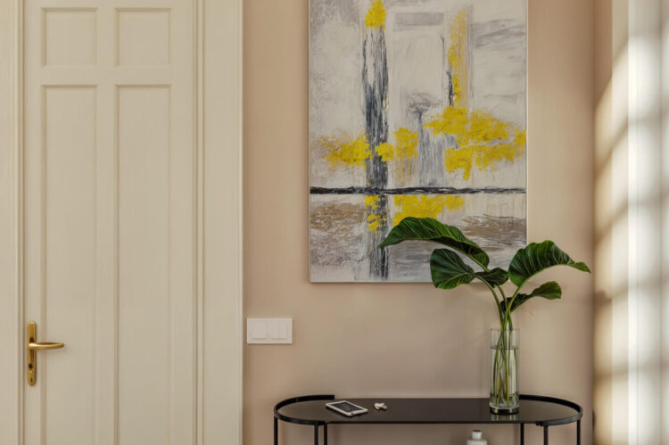 Interior with large abstract painting in yellow and gray, black shelving unit with vases, magazines, and lantern, white door with golden handle, wooden floor, and beige walls.