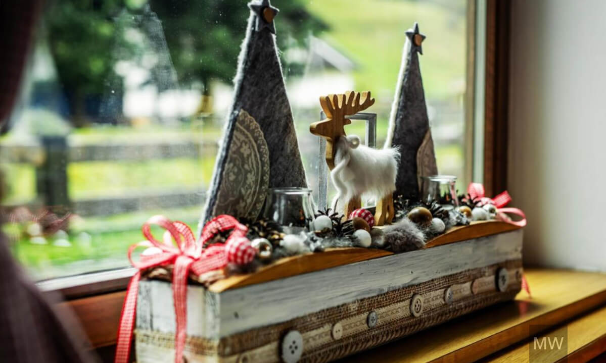 Aranjament decorativ de iarnă cu ren și brazi de fetru pe pervaz. Decor rustic pe pervaz cu figurină de ren și conuri de pin.
