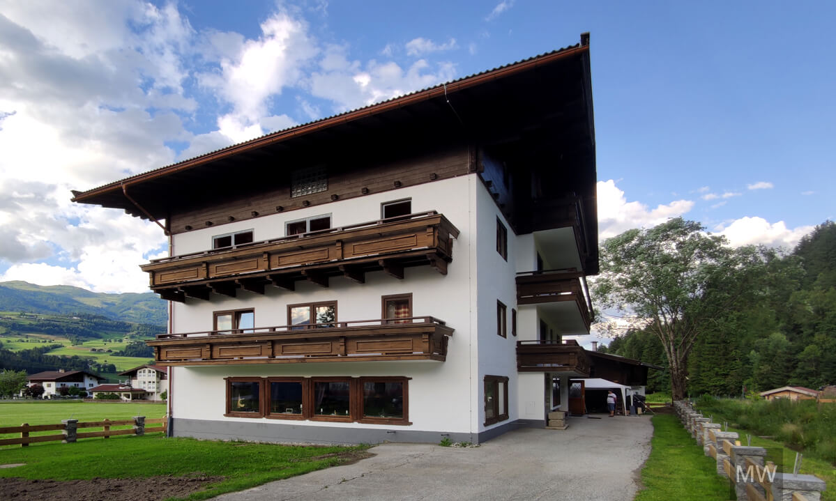 Design interior pensiune rustică cu balcoane din lemn Clădire cu mai multe etaje, cu exterior alb și balcoane din lemn închis, situată pe un fundal de câmpuri verzi și păduri.