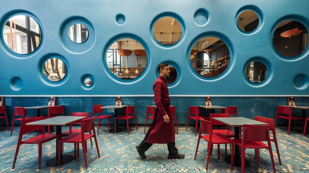Stylish Cafe Design with Blue Wall and Mosaic Tiled Floor | Most Wanted Design Cluj Cafe interior with blue walls, circular mirrors, mosaic floor tiles, red chairs and tables, and a waiter in deep red tones walking across the space.