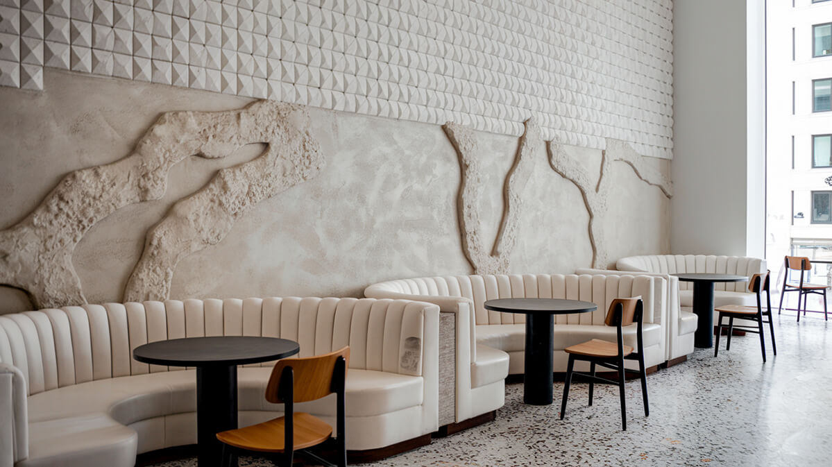 Interior Modern de Cafenea cu Perete Texturat și Design Elegant | Most Wanted Design Cluj Interior modern de cafenea cu perete texturat geometric, canapele albe luxoase, mese cilindrice negre, pardoseală terrazzo alb-gri și vedere panoramică.