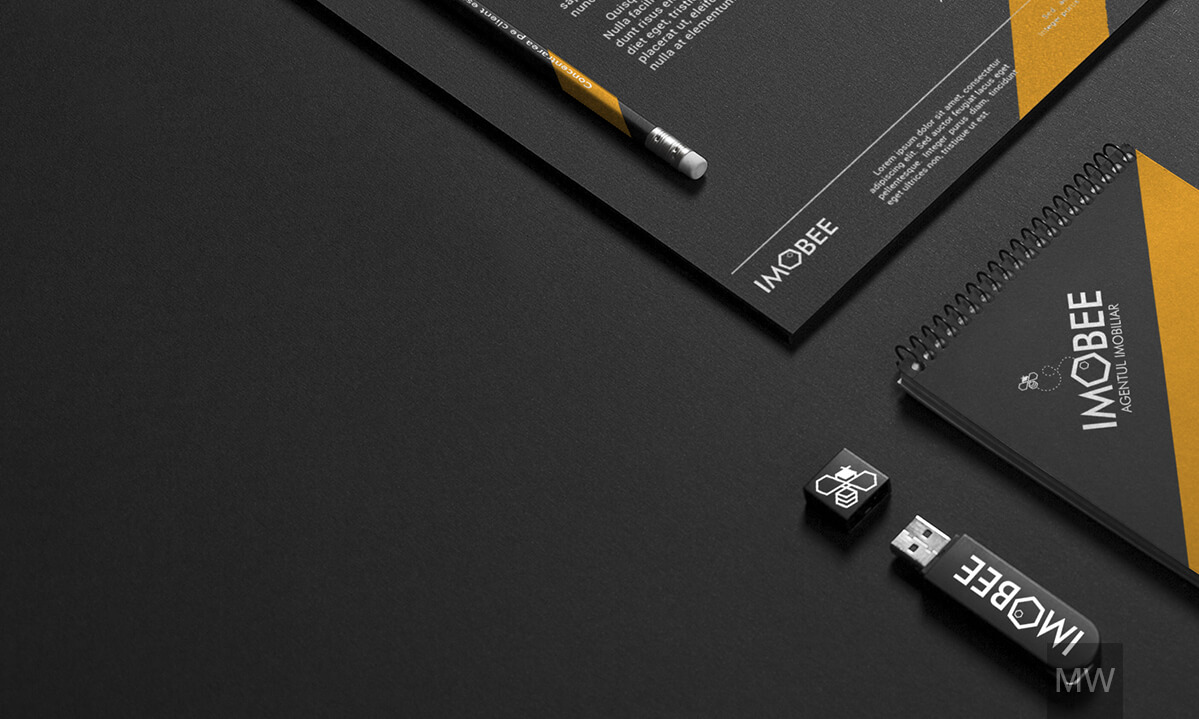 IMOBEE Dark-Themed Branding Setup A dark-themed branding setup for IMOBEE, including a paper with the logo, a pencil, a spiral-bound notebook, and two USB flash drives.
