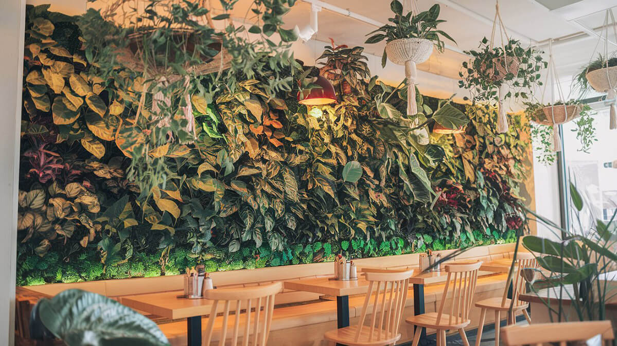 Cafenea vibrantă cu un perete verde impresionant O fotografie a unui interior vibrant de cafenea cu un perete verde spectaculos plin de plante luxuriante. Mobilierul cald din lemn și iluminarea discretă creează o atmosferă primitoare.