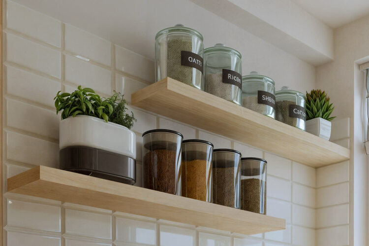 A modern kitchen shelf with labeled transparent jars containing various grains and seeds, set against a white tiled wall.