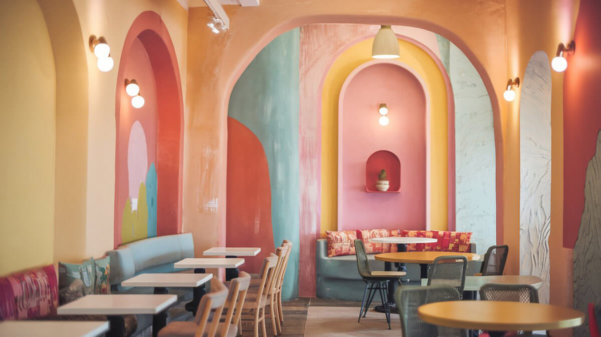 Interior confortabil de cafenea cu paletă de culori pastelate calde O fotografie a unui interior de cafenea confortabilă, cu o paletă de culori pastelate calde. Pereții au un amestec de finisaje netede și vopsea texturată, cu accente de nuanțe vibrante și îndrăznețe.