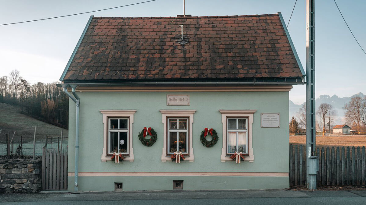 Charming Alpine House with Festive Decor A muted green house with a red-tiled roof in an Austrian alpine village, featuring festive wreaths on two windows.