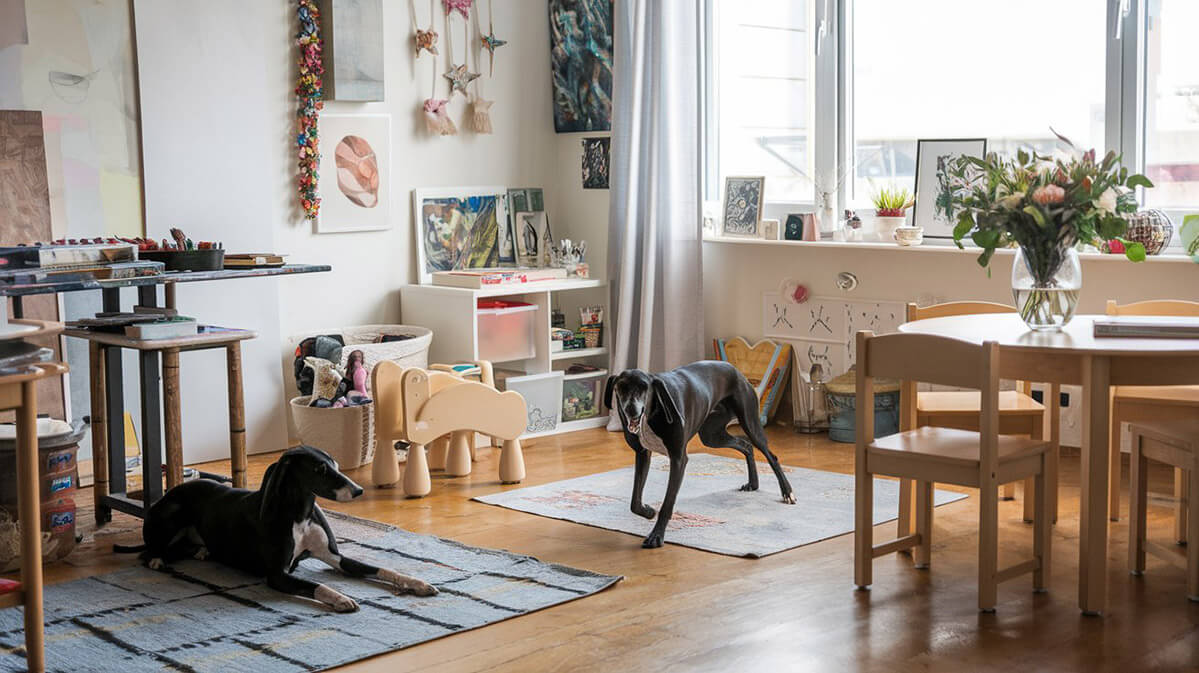 Cozy Artist’s Studio with Playful Dogs An artist's home studio with two black hound-like dogs, one lying on a rug and the other playful.