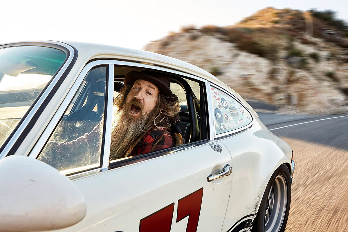 Magnus Walker Driving a White Sports Car Magnus Walker, with long hair, a beard, and a hat, appears excited while seated in a white sports car marked with the number '77,' speeding on a winding road.