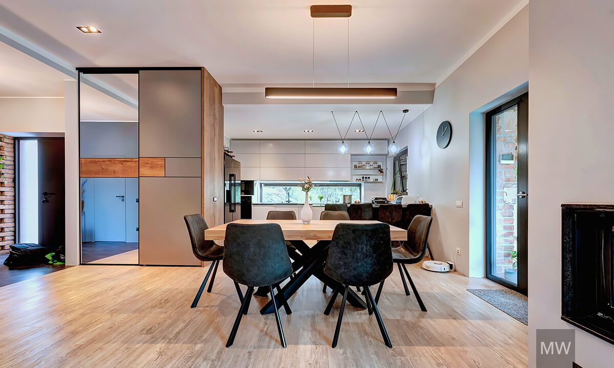 Zonă de dining elegantă cu masă din lemn și accente contemporane Zonă de dining modernă cu masă din lemn, scaune închise la culoare, bucătărie albă în fundal și șemineu pe partea dreaptă.