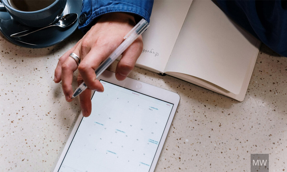 Planificare digitală într-un spațiu de lucru relaxat Mână ținând un stilou digital deasupra unei tablete care afișează un calendar, așezată pe un blat cu suprafață texturată.
