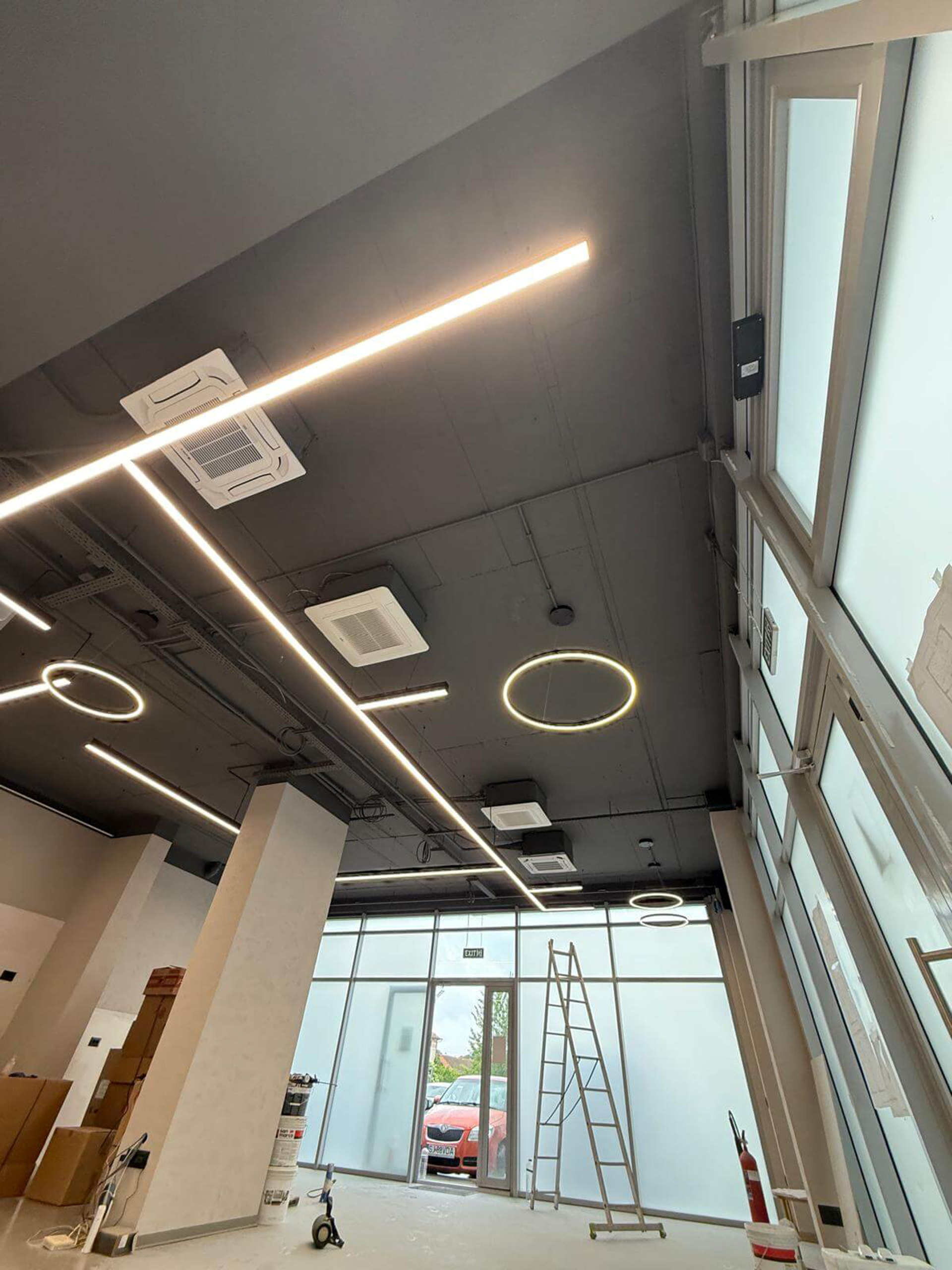 Renovation Scene Featuring Exposed Ceiling and Lighting Design Interior space under renovation with exposed ceiling lights, large windows, a visible red car outside, and construction tools on the floor.