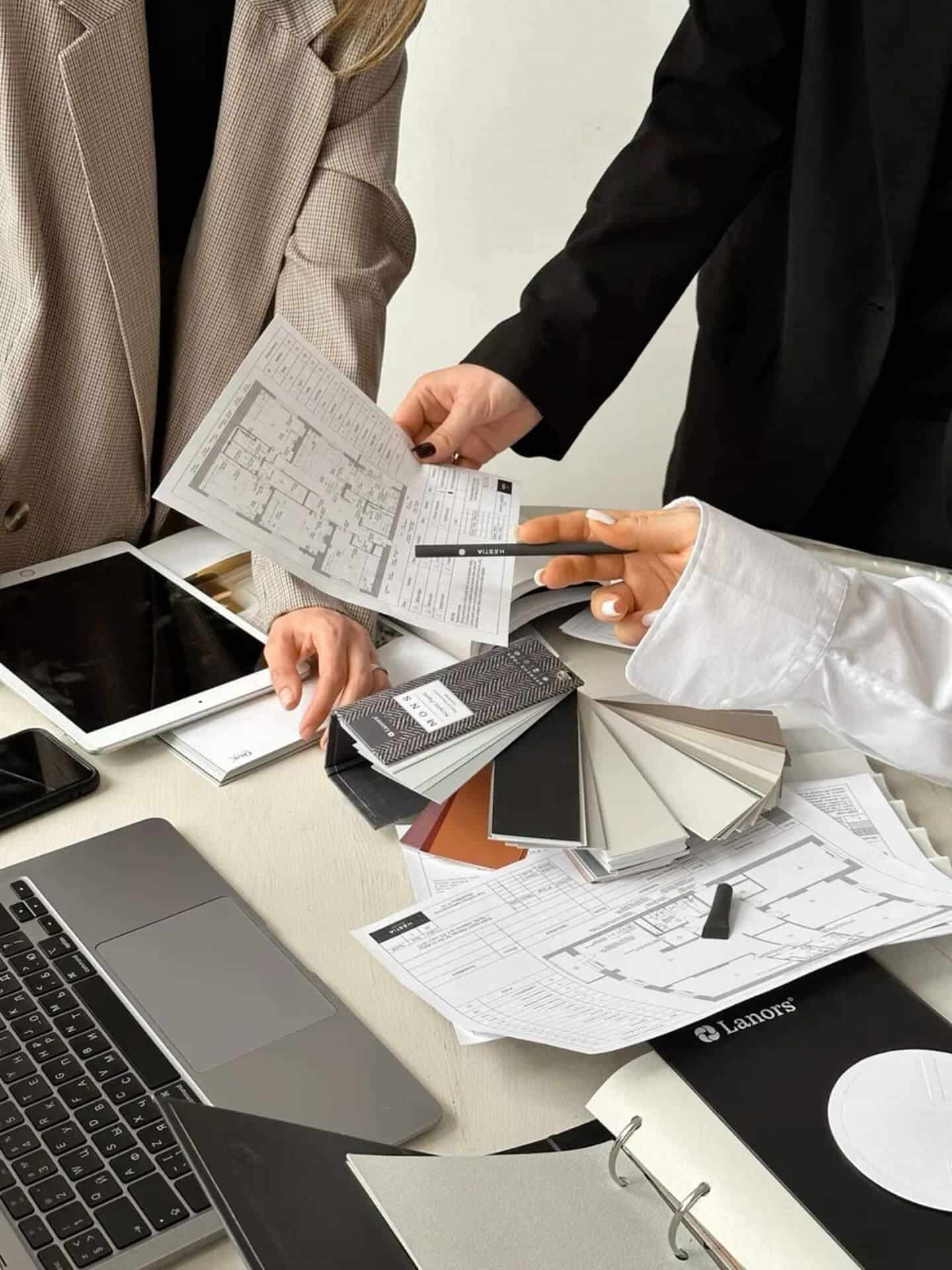 Discuție design interior și arhitectură Două persoane discutând la un birou cu planuri arhitecturale, mostre de materiale și dispozitive electronice, analizând alegeri de design.